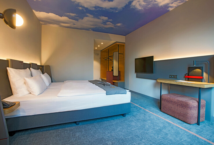 Business Zimmer im ATLANTIC Hotel Airport Bremen Modernes Hotelzimmer mit Doppelbett, Schreibtisch, Flachbild-TV und Himmelmotiv an der Decke im ATLANTIC Hotel Airport.