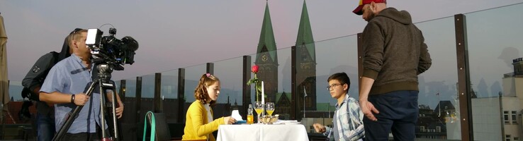 Web-Serie der ATLANTIC Hotels "Stille Post" Paar beim Abendessen auf einer Hotel-Dachterrasse, umgeben von Kamerateam, mit Blick auf Kirchtürme im Hintergrund.