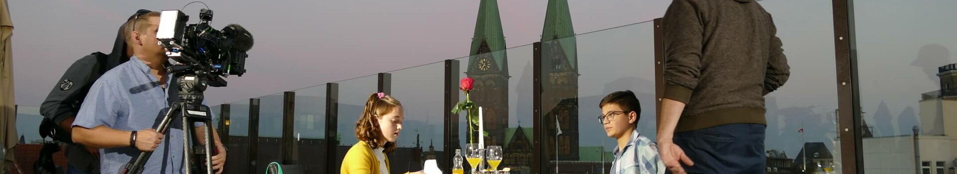 Paar beim Abendessen auf einer Hotel-Dachterrasse, umgeben von Kamerateam, mit Blick auf Kirchtürme im Hintergrund.