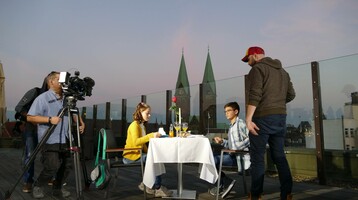 Paar beim Abendessen auf einer Hotel-Dachterrasse, umgeben von Kamerateam, mit Blick auf Kirchtürme im Hintergrund.