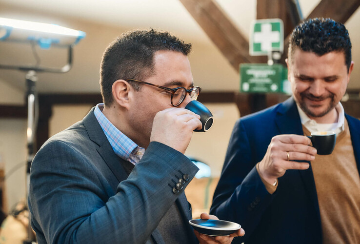 Zwei Männer in Anzügen trinken Kaffee und lächeln in einem gemütlichen Raum.