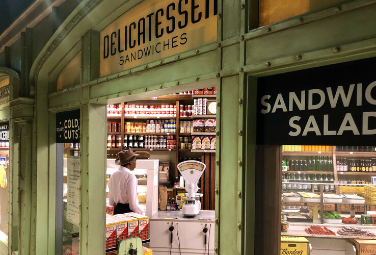 Ein Delikatessengeschäft mit Sandwiches und Salaten, Verkäufer hinter der Theke mit Waage und Regalen voller Produkte.