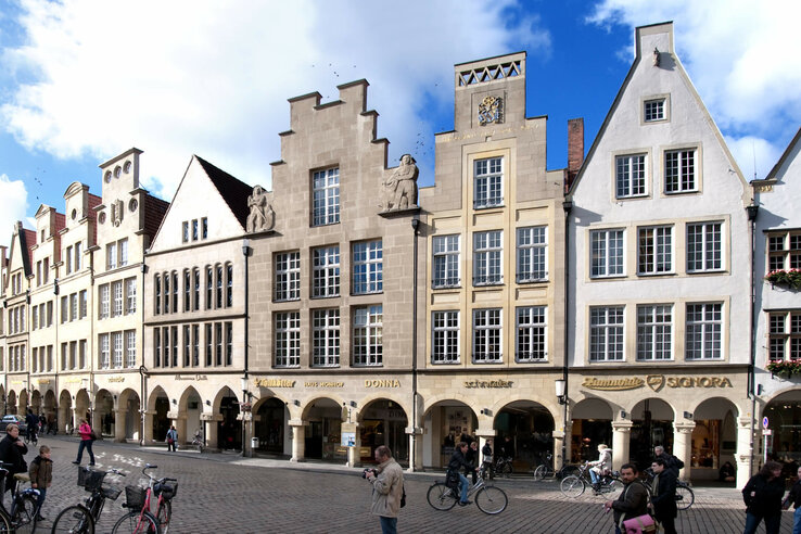 Historic facades on Prinzipalmarkt in Münster under a blue sky, with people and bicycles on the cobblestones.