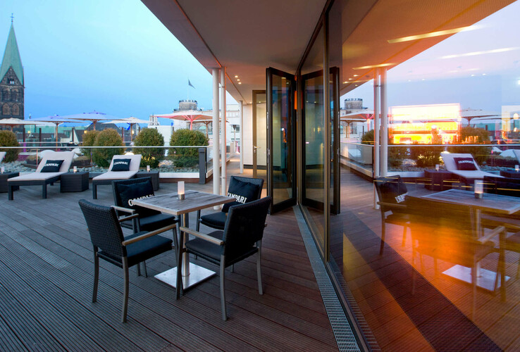 Hotelterrasse mit Sitzgruppen, Liegen und Blick auf historische Kirchtürme bei Abenddämmerung.