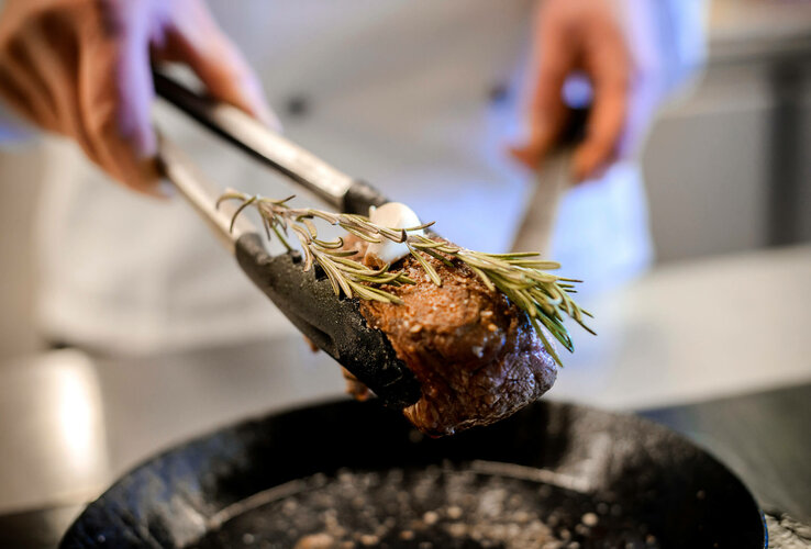 Koch hält Steak mit Rosmarin und Knoblauch über einer Pfanne im ATLANTIC Hotel Kiel.