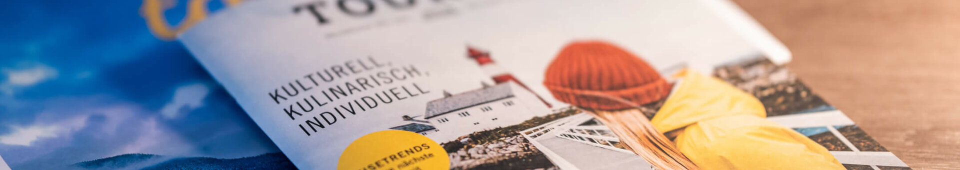 Two travel magazines on a wooden table, one showing a person in a yellow jacket in front of a lighthouse.