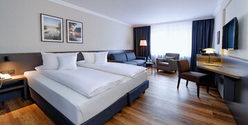 Modernes Hotelzimmer mit Doppelbett, Sofa, Sessel, Schreibtisch, TV und großem Fenster mit Vorhängen.