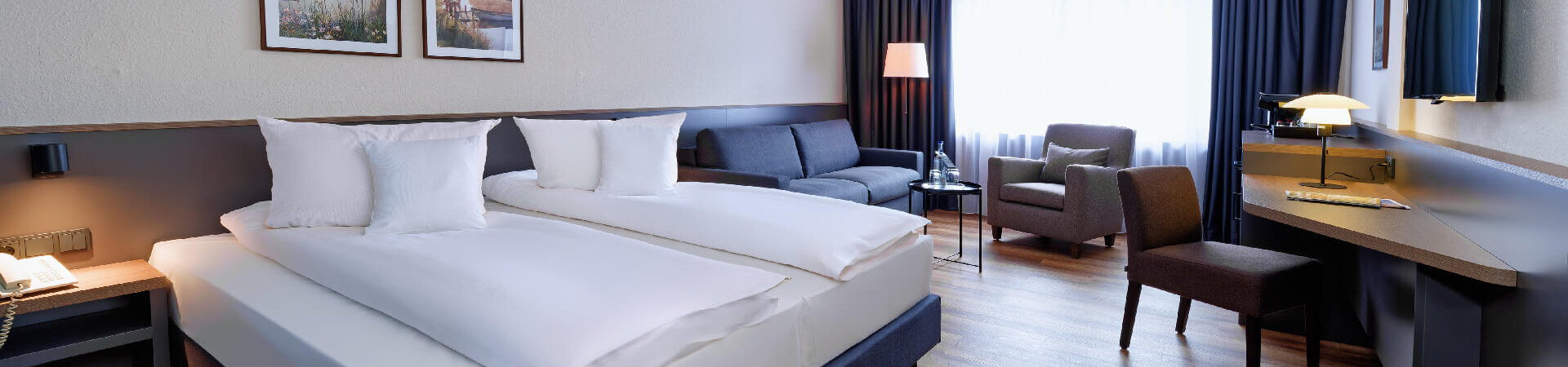 Deluxe Zimmer im ATLANTIC Hotel Landgut Horn Modernes Hotelzimmer mit Doppelbett, Sofa, Sessel, Schreibtisch, TV und großem Fenster mit Vorhängen.