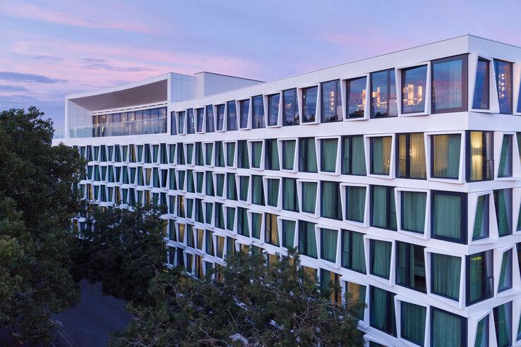 Modern hotel building with large windows and green curtains, surrounded by trees, at sunset.