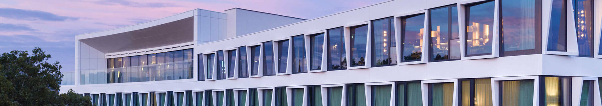 Modernes Hotelgebäude mit großen Fenstern und grünen Vorhängen, umgeben von Bäumen, bei Sonnenuntergang.