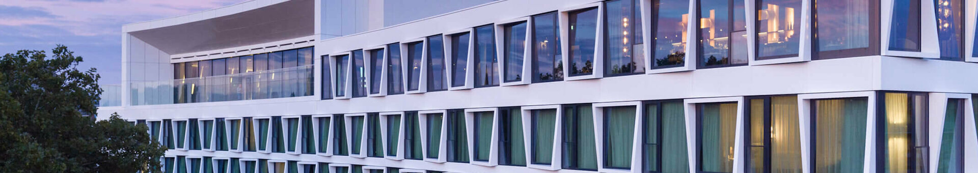 Aussenfassade ATLANTIC Hotel Münster Modernes Hotelgebäude mit großen Fenstern und grünen Vorhängen, umgeben von Bäumen, bei Sonnenuntergang.