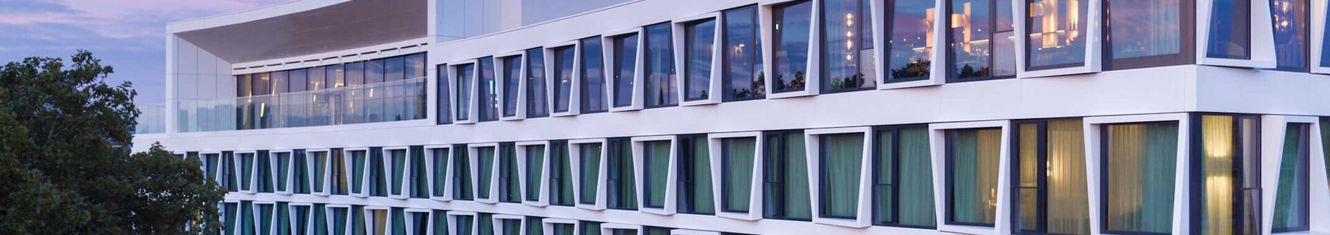 Modern hotel building with large windows and green curtains, surrounded by trees, at sunset.