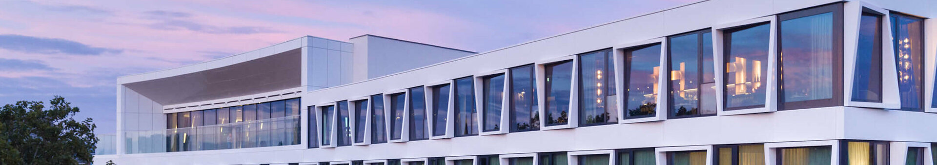 Exterior Facade from ATLANTIC Hotel Münster Modern hotel building with large windows and green curtains, surrounded by trees, at sunset.