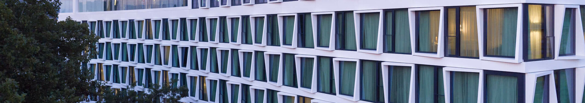 Exterior Facade from ATLANTIC Hotel Münster Modern hotel building with large windows and green curtains, surrounded by trees, at sunset.