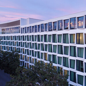 Modernes Hotelgebäude mit großen Fenstern und grünen Vorhängen, umgeben von Bäumen, bei Sonnenuntergang.