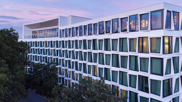 Modernes Hotelgebäude mit großen Fenstern und grünen Vorhängen, umgeben von Bäumen, bei Sonnenuntergang.