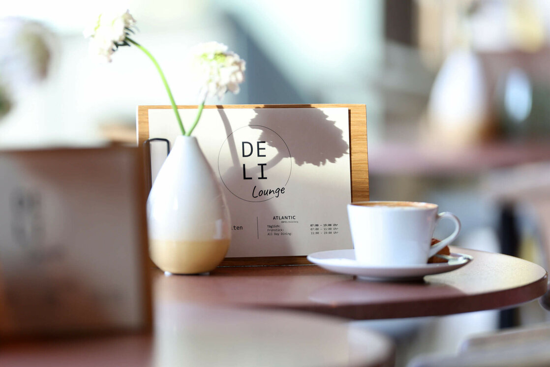 Hausgerösteter Barista Kaffee in der Deli Lounge im ATLANTIC Hotel Heidelberg Kaffeetasse und Vase auf Tisch in der DELI Lounge des ATLANTIC Hotel Heidelberg, mit Menü im Hintergrund.