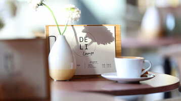 Hausgerösteter Barista Kaffee in der Deli Lounge im ATLANTIC Hotel Heidelberg Kaffeetasse und Vase auf Tisch in der DELI Lounge des ATLANTIC Hotel Heidelberg, mit Menü im Hintergrund.
