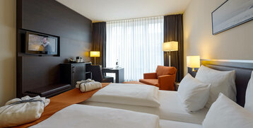 Helles Hotelzimmer mit Doppelbett, TV, Schreibtisch, Sessel und großen Fenstern im ATLANTIC Hotel Lübeck.