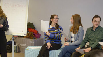 The trainees sit in a large circle of chairs while planning the trainee day.