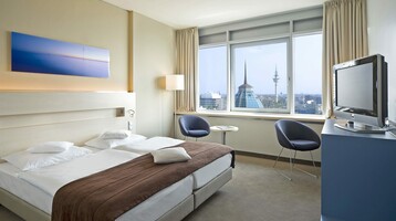 Bright hotel room with double bed, two armchairs, TV and large window with a view of Bremerhaven's skyline.