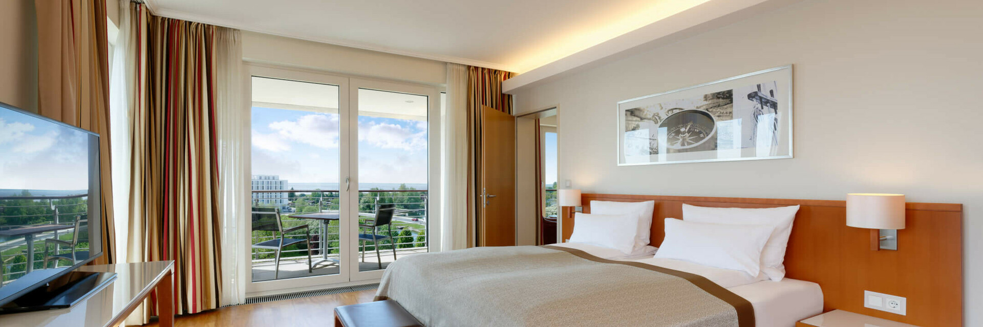Helles Hotelzimmer mit Doppelbett, Holzboden, TV, Balkon mit Stühlen und Blick auf grüne Landschaft.