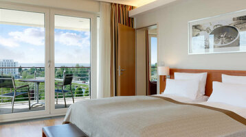 Helles Hotelzimmer mit Doppelbett, Holzboden, TV, Balkon mit Stühlen und Blick auf grüne Landschaft.