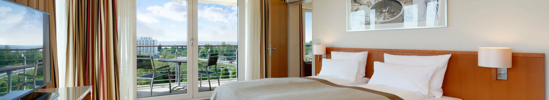 Übernachtungsangebot 5 für 4 Helles Hotelzimmer mit Doppelbett, Holzboden, TV, Balkon mit Stühlen und Blick auf grüne Landschaft.