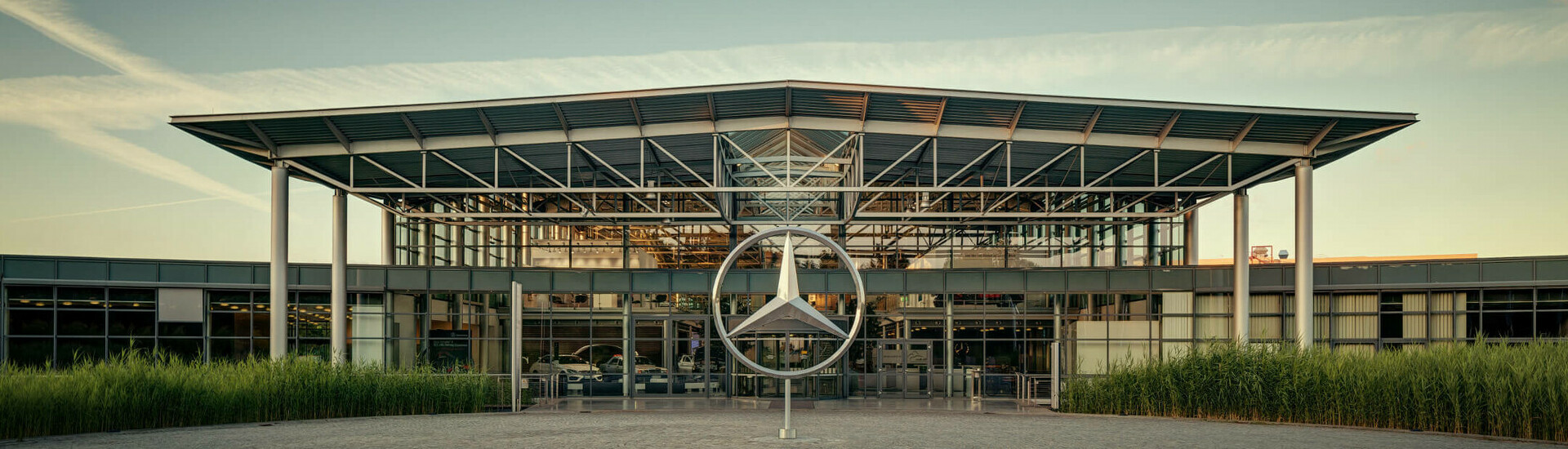 Modernes Gebäude mit Glasfassade und großem Mercedes-Logo im Vordergrund, umgeben von grünen Pflanzen.