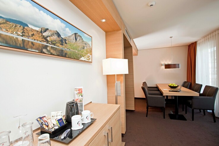 Helles Hotelzimmer mit Esstisch, Stühlen, Kaffeestation und Landschaftsbild an der Wand.