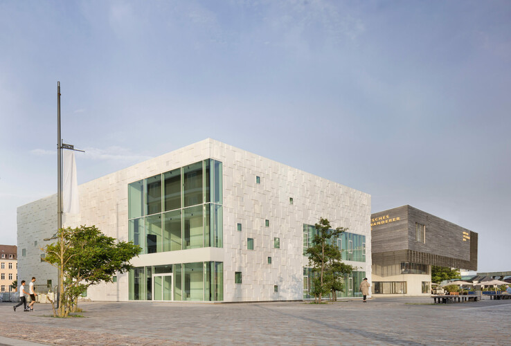 Modernes Gebäude mit Glasfassade und weißen Wänden, umgeben von einem gepflasterten Platz, bei klarem Himmel.