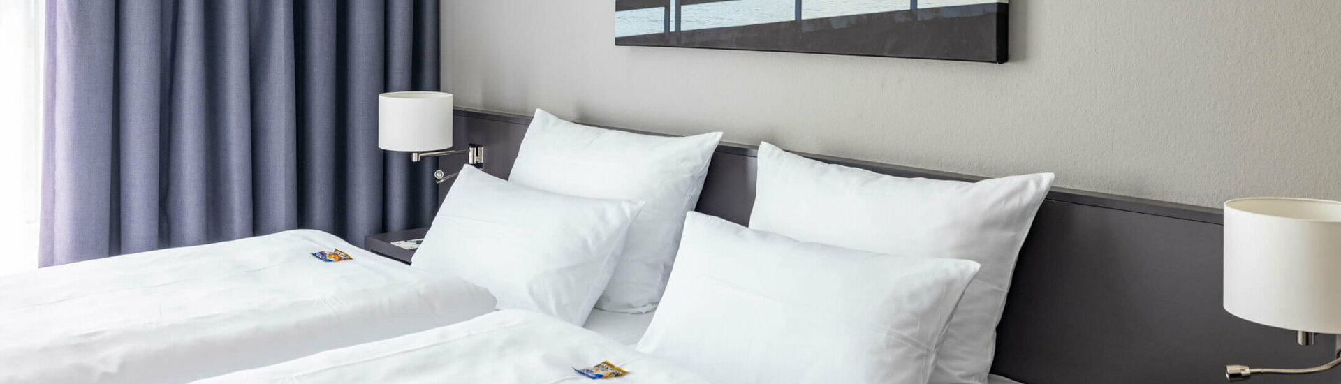 ATLANTIC Hotel Vegesack Comfort Doppelzimmer Details Modernes Hotelzimmer mit zwei Einzelbetten, blauen Vorhängen und maritimem Wandbild.