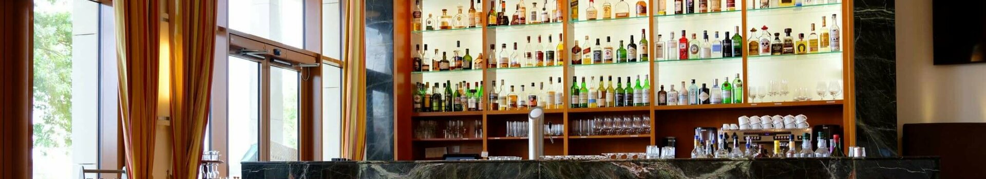 ATLANTIC Hotel Wilhelmshaven Seven C´s Bar Elegant hotel bar with illuminated shelves full of spirits, leather armchairs and large windows with curtains.