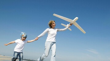 Eine Frau und ein Kind laufen am Strand, halten Hände und tragen weiße Kleidung, während sie ein Spielzeugflugzeug fliegen.