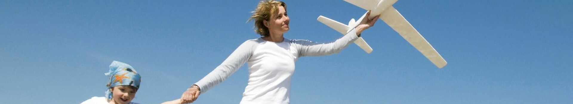 Eine Frau und ein Kind laufen am Strand, halten Hände und tragen weiße Kleidung, während sie ein Spielzeugflugzeug fliegen.