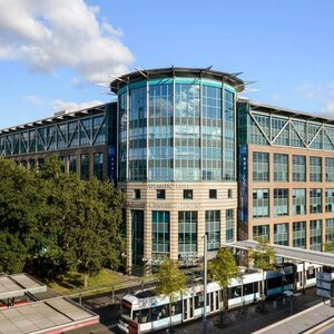Modern hotel building with glass façade, surrounded by trees, close to a streetcar stop.