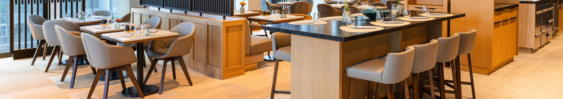 A bright, modern dining area at the ATLANTIC Hotel Frankfurt, featuring wood and natural tones. Several tables and upholstered chairs are arranged in front of large windows. In the centre is a large high table with seating. Striking, ring-shaped pendant lights and high-quality materials create an elegant and inviting atmosphere.
