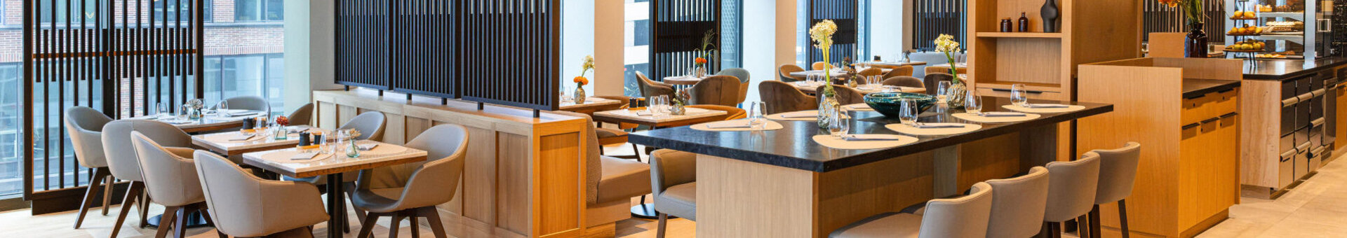 Heller, modern gestalteter Gastronomiebereich im ATLANTIC Hotel Frankfurt mit Holz- und Naturtönen. Mehrere Tische und gepolsterte Stühle stehen vor großen Fenstern. In der Mitte befindet sich ein großer Hochtisch mit Sitzgelegenheiten. Auffällige, ringförmige Pendelleuchten und hochwertige Materialien schaffen eine elegante und einladende Atmosphäre.