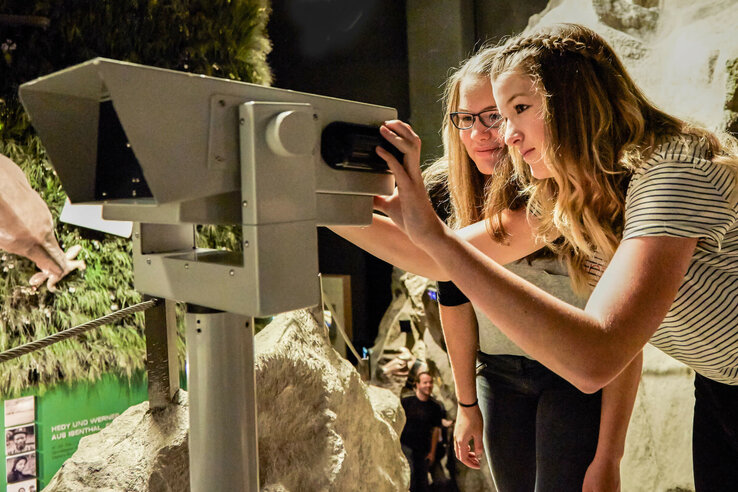 Two women look through binoculars at an exhibition in a museum.