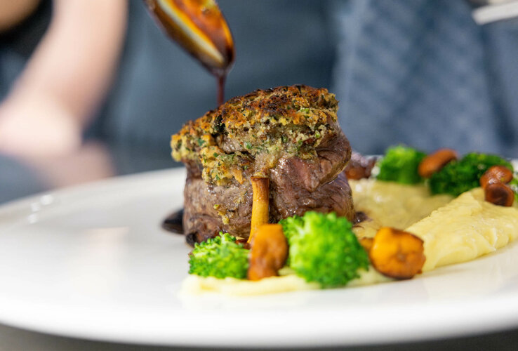 Gourmetgericht mit Fleisch, Brokkoli, Pilzen und Püree auf weißem Teller, mit dunkler Sauce garniert.
