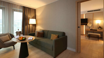 Modernes Hotelzimmer mit gemütlicher Sitzecke, Sofa, Tisch mit Obst, Lampe und Blick ins angrenzende Zimmer mit TV.