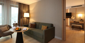 Modernes Hotelzimmer mit gemütlicher Sitzecke, Sofa, Tisch mit Obst, Lampe und Blick ins angrenzende Zimmer mit TV.
