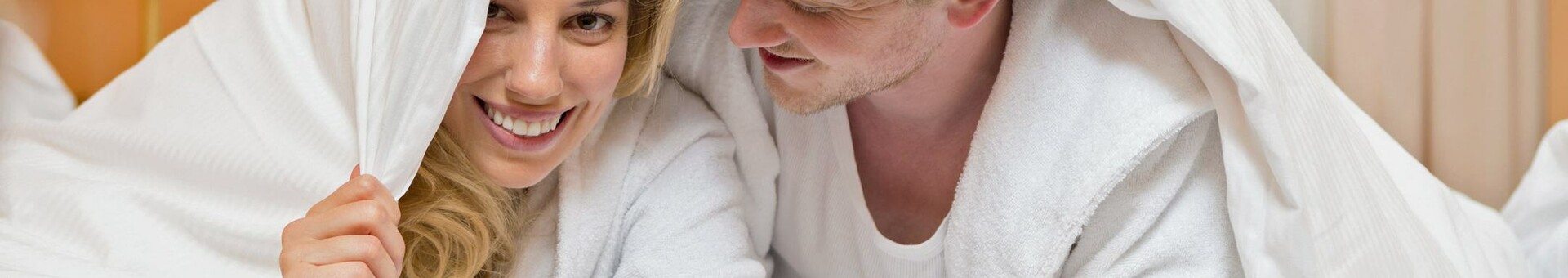 Ein lächelndes Paar in Bademänteln liegt spielerisch unter einer Decke.
