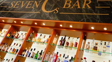 Regal mit beleuchteten Spirituosenflaschen unter dem Schild "Seven Cs Bar" auf dunklem Hintergrund.