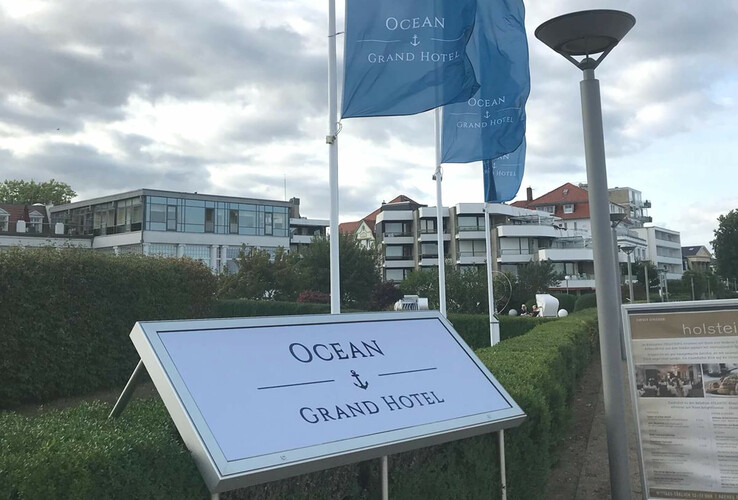 Hotelgebäude mit modernen Balkonen, umgeben von gepflegten Hecken, blauen Fahnen und einem Schild "Ocean Grand Hotel".