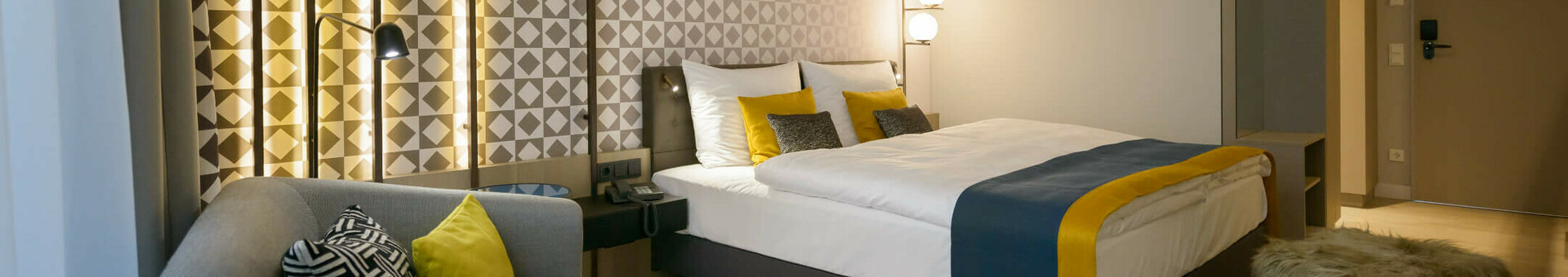 ATLANTIC Hotel Münster Zimmer Modernes Hotelzimmer mit Doppelbett, stilvoller Beleuchtung, grauem Sofa und gemusterten Kissen im ATLANTIC Hotel Münster.