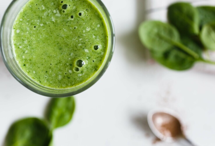 Green smoothie in a glass, next to fresh spinach leaves and a spoon with powder on a white background.