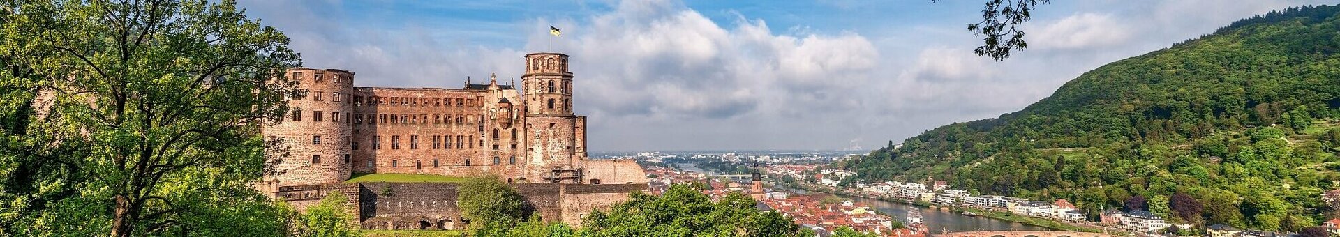 ATLANTIC Hotel Heidelberg Rahmenprogramm Header Blick auf ein historisches Schloss und eine Stadt am Fluss, umgeben von grünen Hügeln und Bäumen unter blauem Himmel.