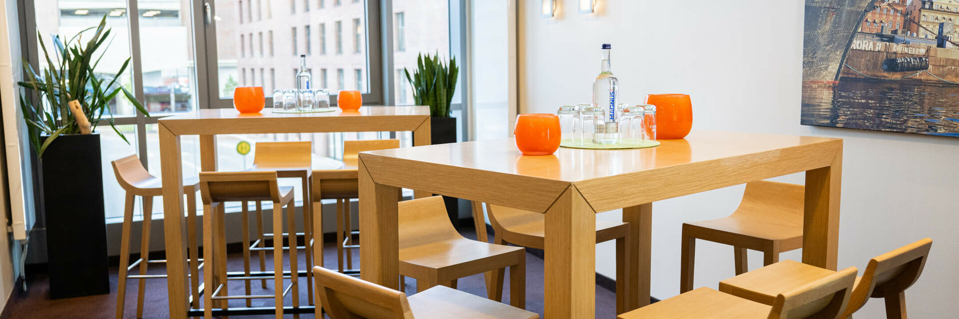 Moderner Lounge-Bereich mit hohen Holztischen, Hockern, Pflanzen und Blick auf die Stadt durch große Fenster.