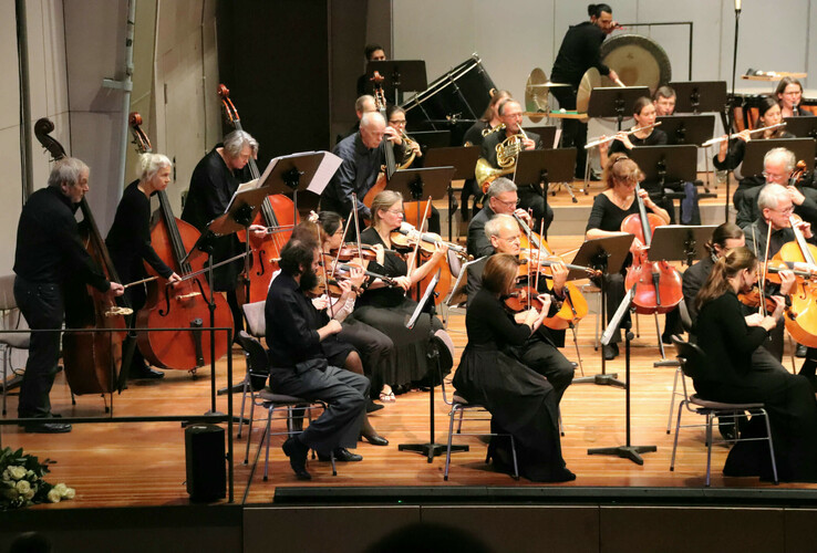 Ein Orchester spielt auf einer Bühne, Musiker mit Streich- und Blasinstrumenten, konzentriert und elegant gekleidet.